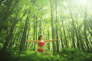 A young woman practices yoga in a sunlit outdoor setting during a forest bathing session surrounded by nature.