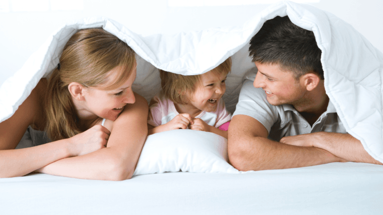 A family—a daughter between her parents—lying under a weighted blanket, smiling and enjoying a moment of happiness and comfort.