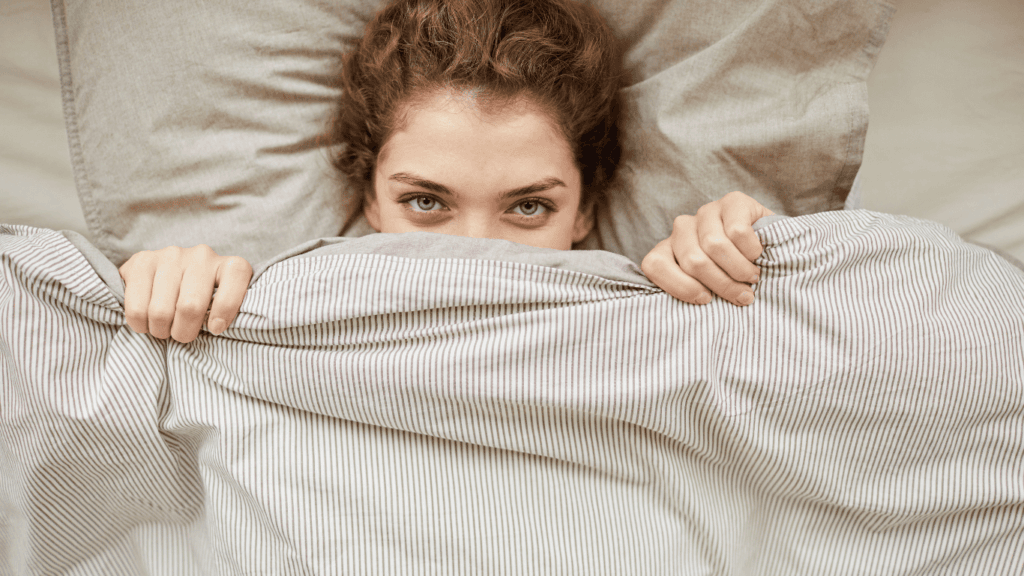 A young woman lying comfortably in bed under a neatly spread weighted blanket, gazing contentedly into the distance.